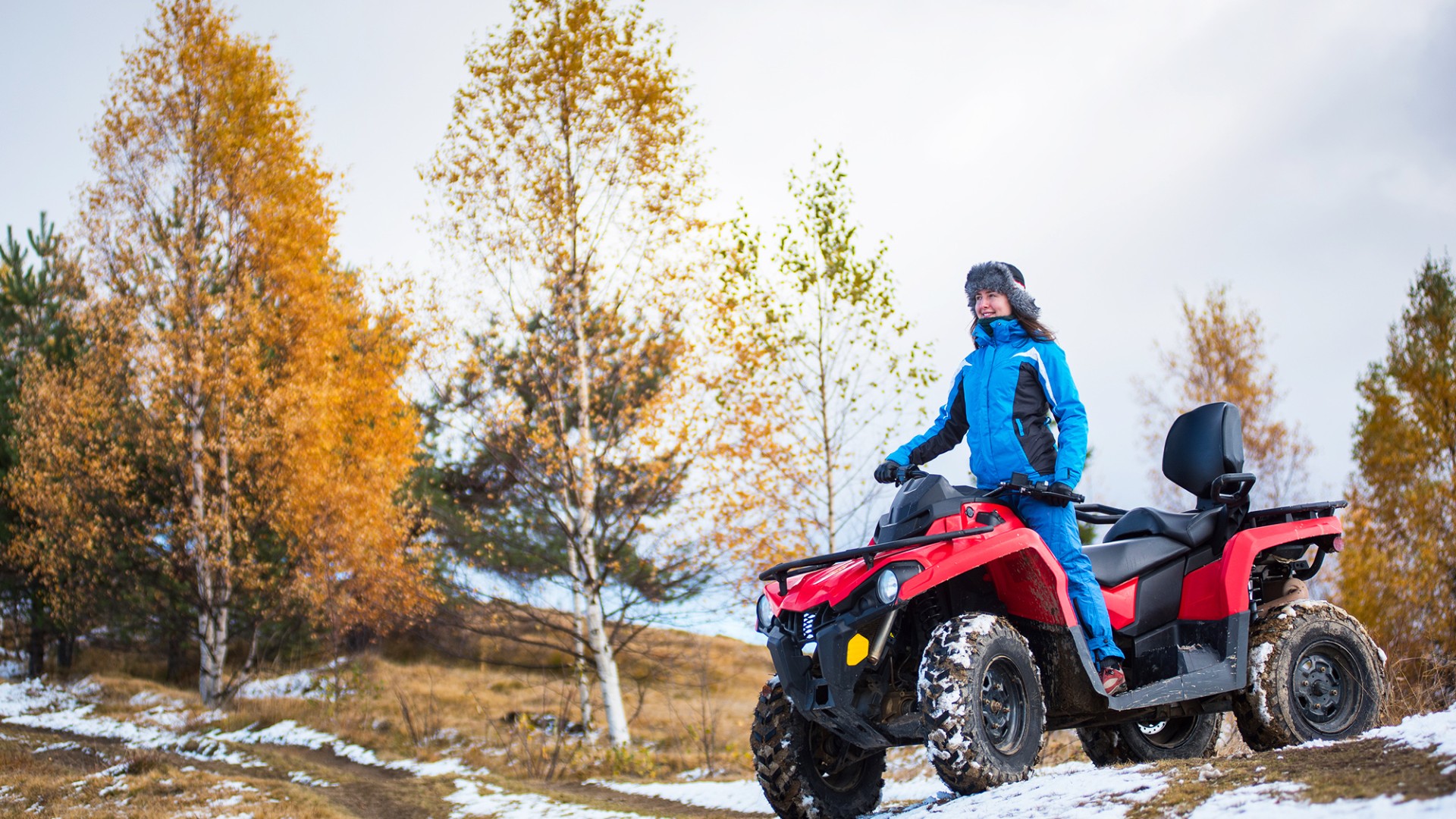 Happy woman in blue winter clothes riding on a red quadbike atv on snow-covered hill against autumn nature on the background with copy space