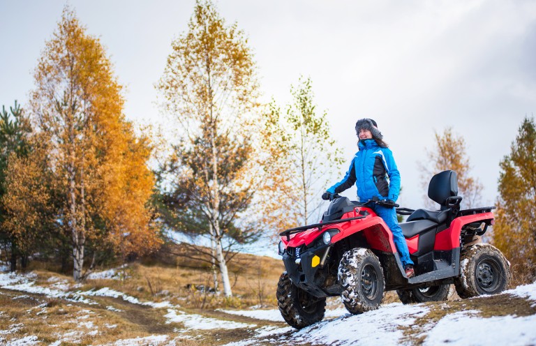 Happy woman in blue winter clothes riding on a red quadbike atv on snow-covered hill against autumn nature on the background with copy space