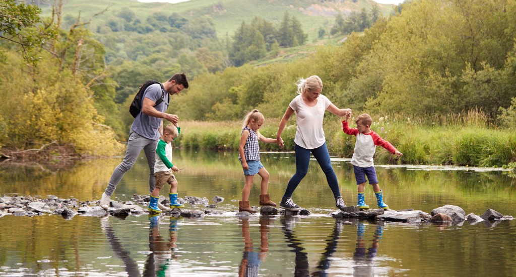 Family Crossing River Whilst Hiking In UK Lake District