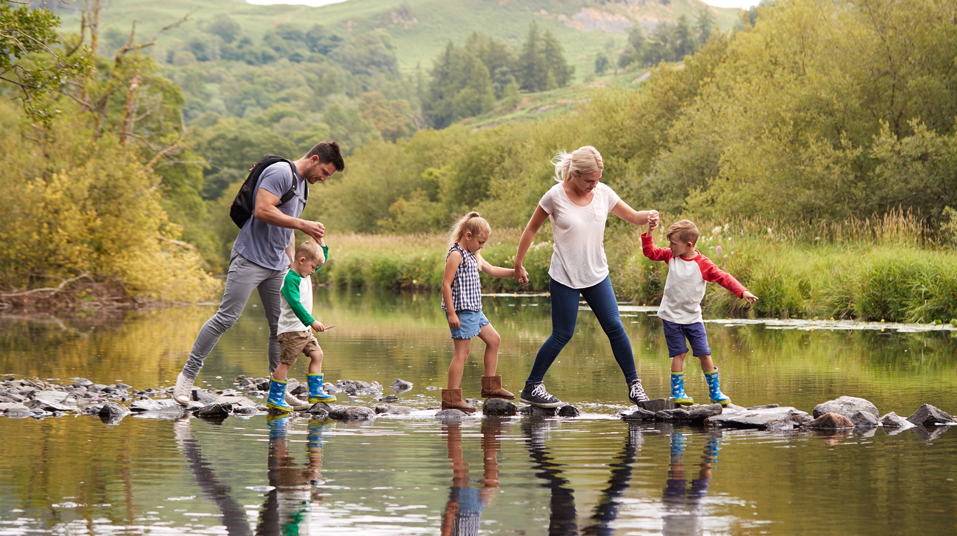 Family Crossing River Whilst Hiking In UK Lake District